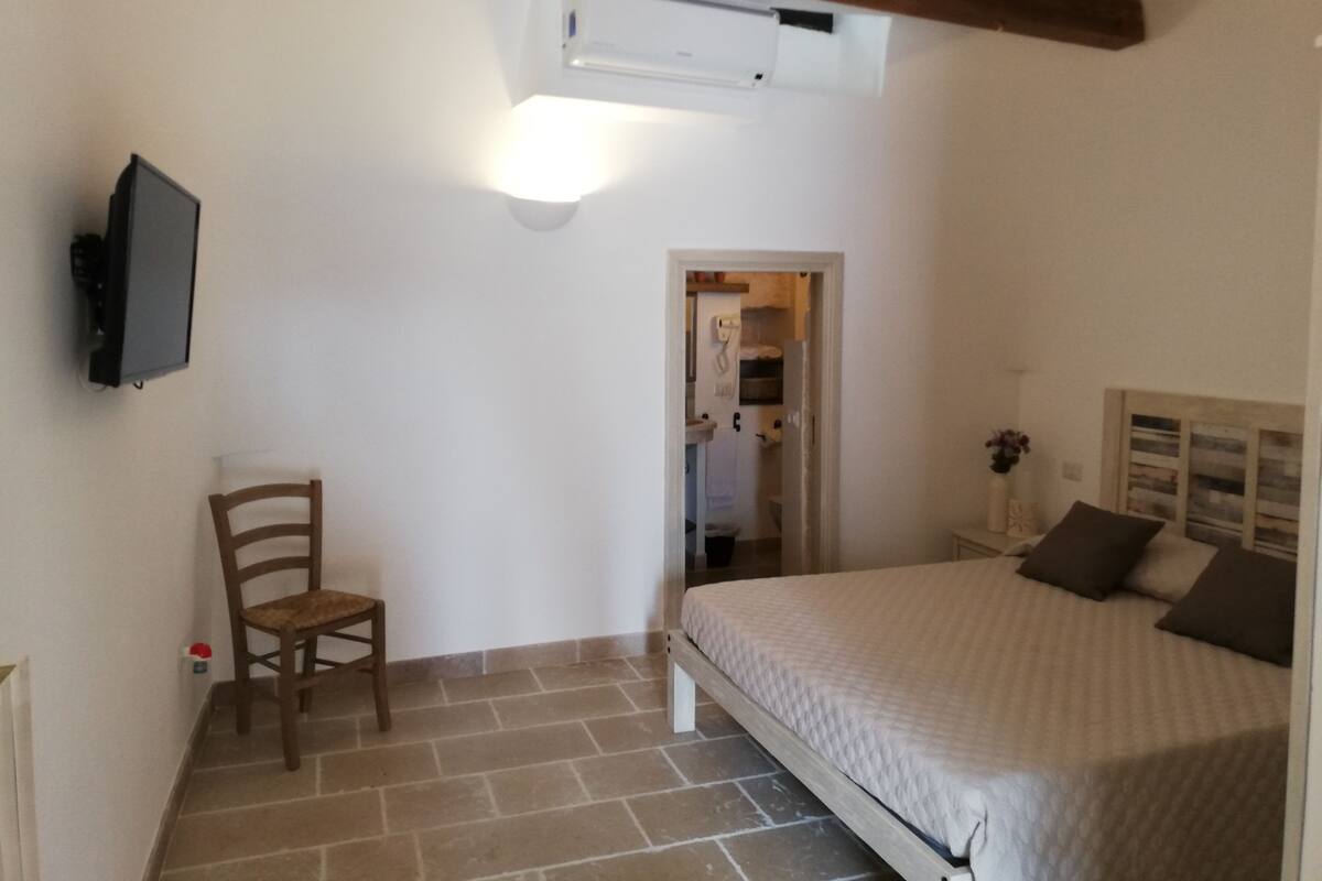 A bedroom with a bed, a chair, a television, and a bathroom visible through an open door.