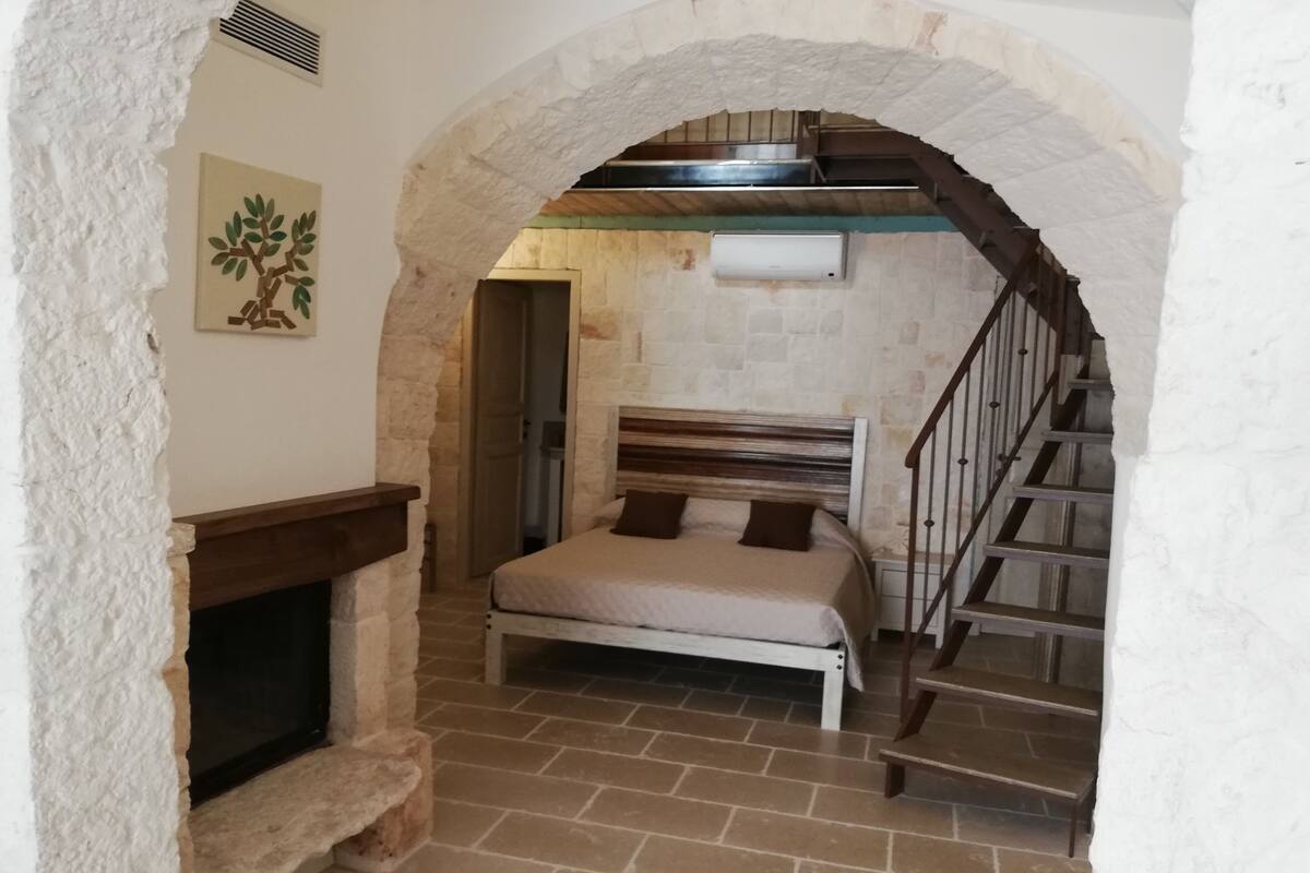 A bedroom with a stone fireplace, a bed with a headboard, and a staircase leading to an upper level.