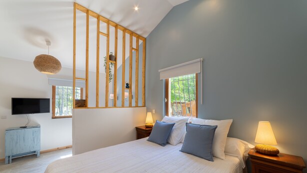 Une chambre avec un grand lit, deux tables de chevet avec des lampes, un téléviseur posé sur un meuble et une fenêtre donnant sur un paysage verdoyant.