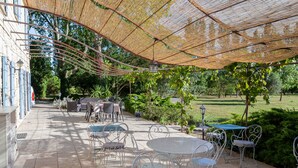 Une terrasse extérieure avec des chaises et des tables en métal, une pergola offrant de l’ombre et une vue sur un jardin et des arbres.