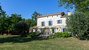 Une maison en pierre à deux étages, avec des volets bleus, entourée de verdure et surplombée d’un ciel bleu azur.