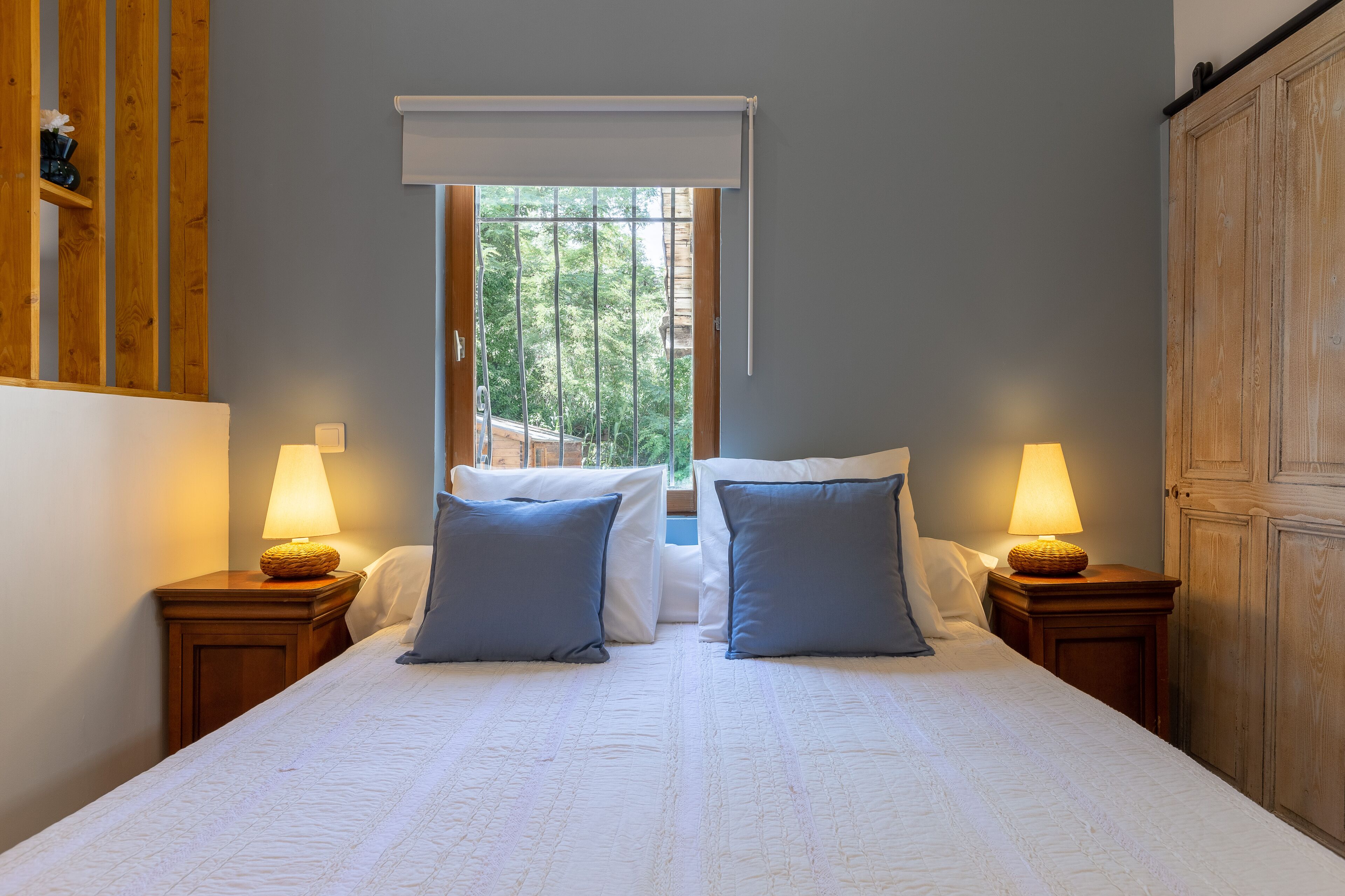 A double bed with two pillows, a window with a view of trees, and two bedside lamps.