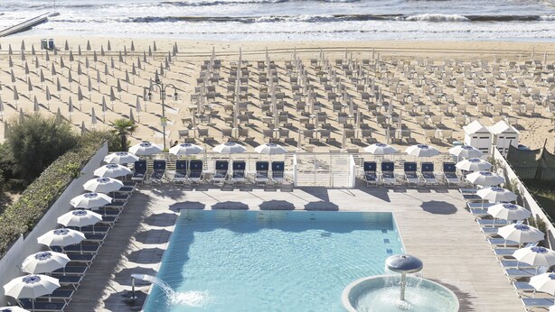 Un área de piscina con sillones de descanso y sombrillas, una playa con filas de sombrillas, y una vista despejada del océano.