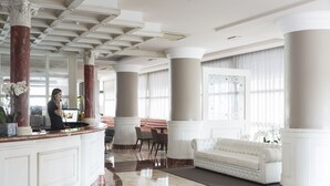 Un vestíbulo amplio con piso de mármol, un mostrador de recepción blanco, un sofá blanco y ventanas grandes con cortinas.