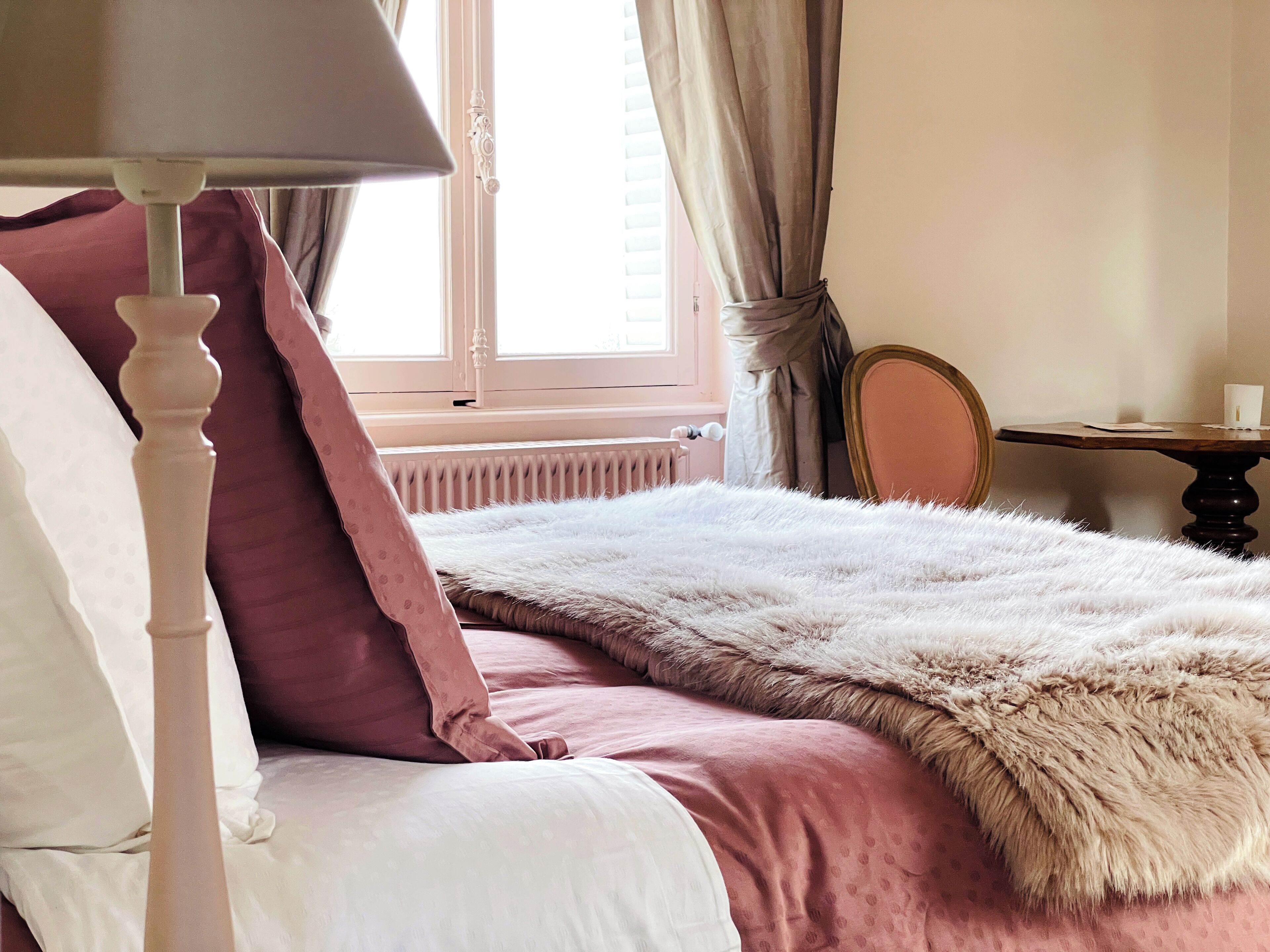 A bedroom with a bed, a bedside lamp, a window with curtains, a radiator, a mirror, and a small table with a cup.