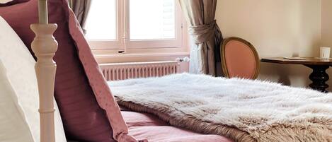 A bedroom with a bed, a bedside lamp, a window with curtains, a radiator, a mirror, and a small table with a cup.