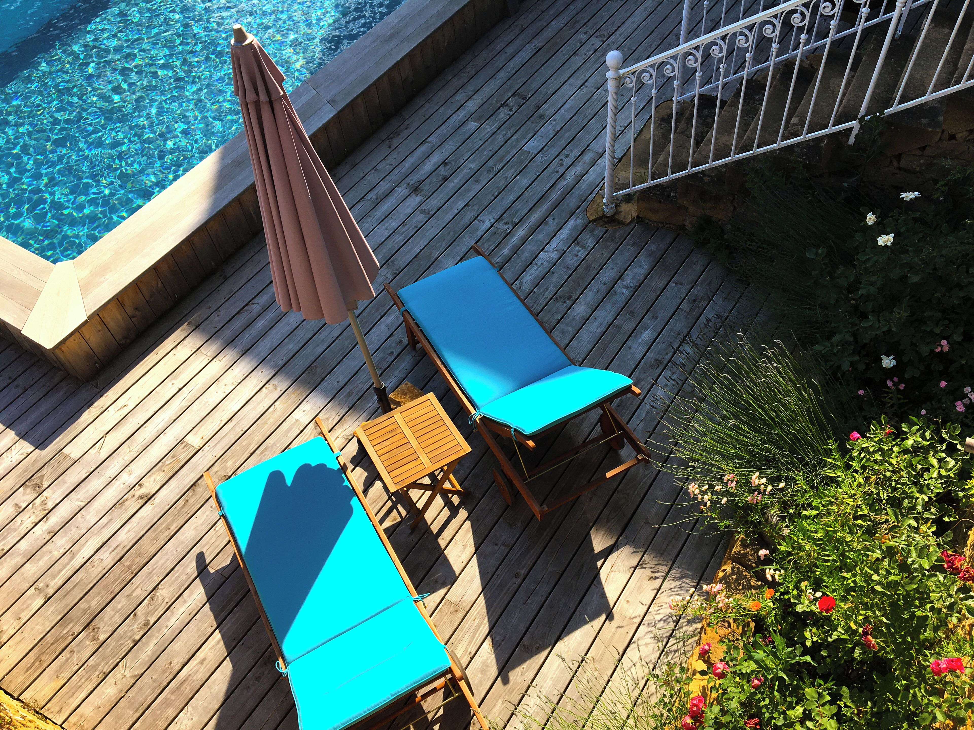 A wooden deck with two blue lounge chairs and a small table, overlooking a swimming pool.