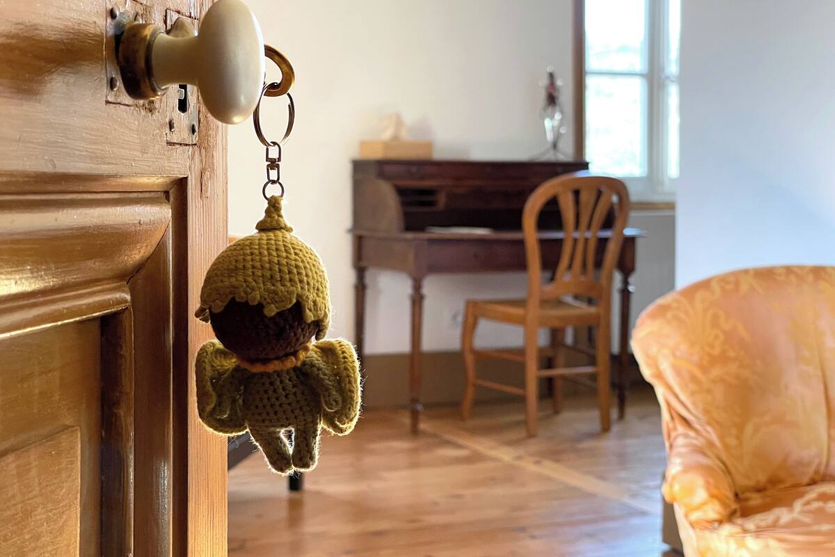 Une porte en bois avec un trou de serrure et une sonnette. Au fond, on aperçoit une chaise en bois et une petite table avec une lampe.