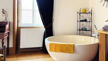 A bathroom with a large freestanding bathtub, a radiator, a wooden floor, and a window with curtains.