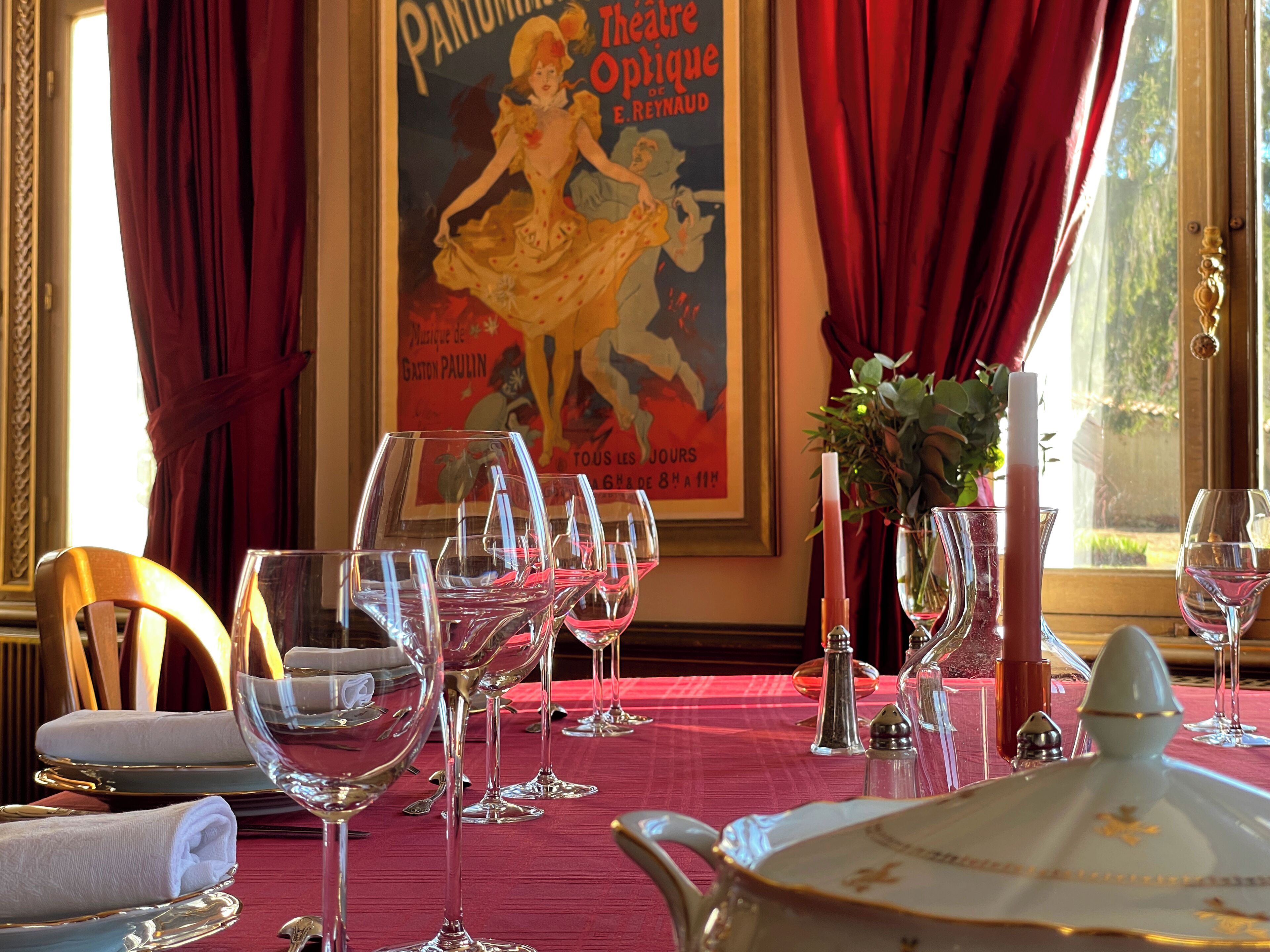 Une table à manger dressée avec des verres à vin, une carafe et une assiette décorative, avec une affiche et des rideaux en arrière-plan.