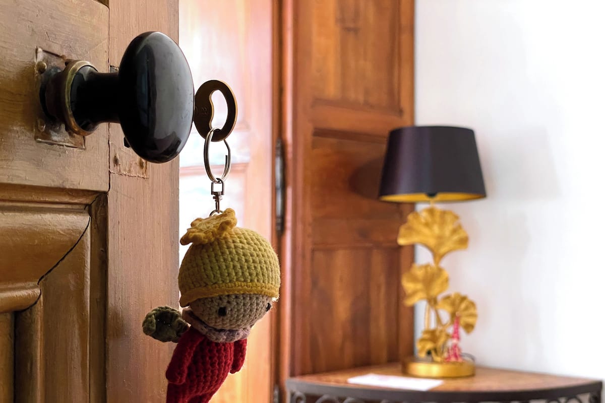 Une porte en bois avec un trou de serrure, un porte-clés orné d’une figurine tricotée et une lampe posée sur une table à l’arrière-plan.