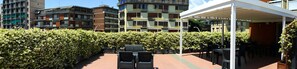 A rooftop patio with outdoor seating, surrounded by a dense hedge and modern buildings in the background. - Terrazza Belfiore (Florence)