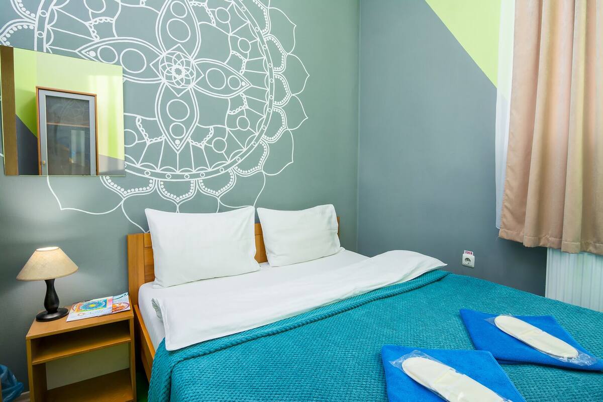 A hotel room with a bed, a nightstand, a lamp, a wall with a decorative pattern, and a window with curtains.