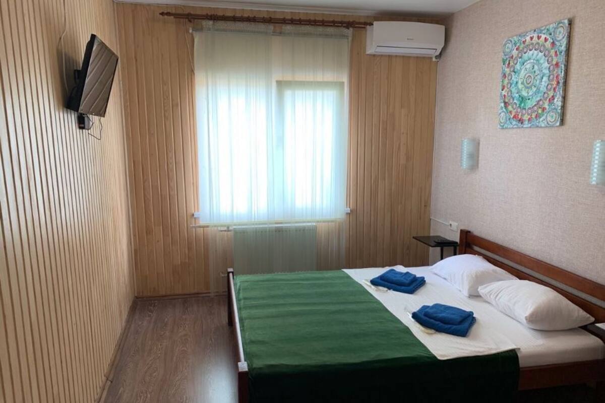 A hotel room with a wooden paneled wall, a bed with a green bedspread and blue towels, a window with curtains, an air conditioning unit, and a wall-mounted TV.