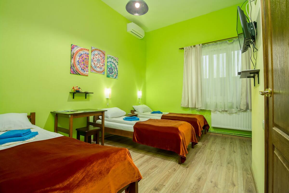 A hotel room with two beds, a desk, and a chair. There are colorful wall decorations and a window with curtains.