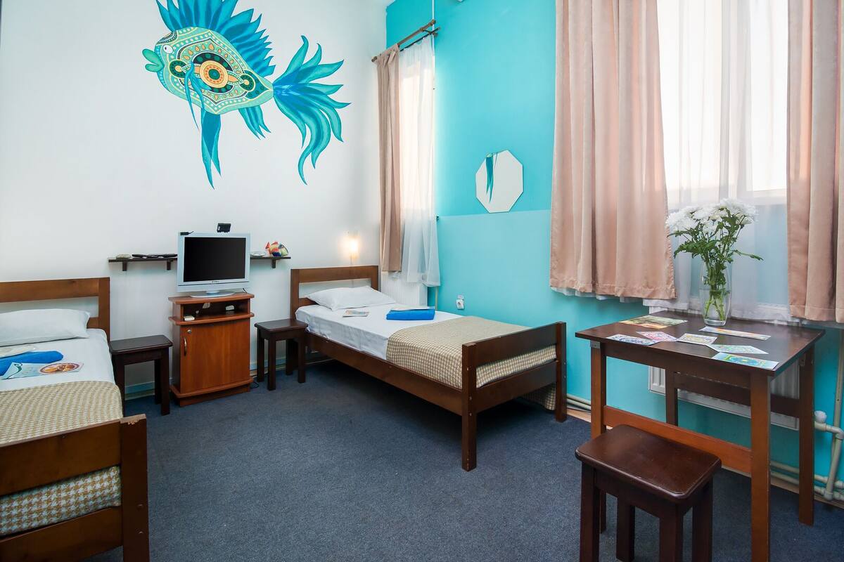 A hotel room with two single beds, a desk with a chair, a television, and a window with curtains.