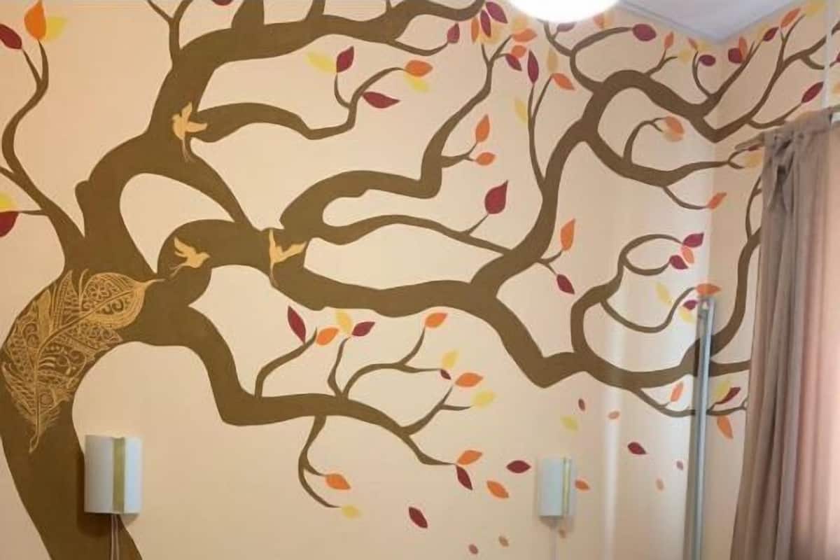 A hotel room with a bed, a nightstand, a lamp, and a wall mural of a tree with leaves.