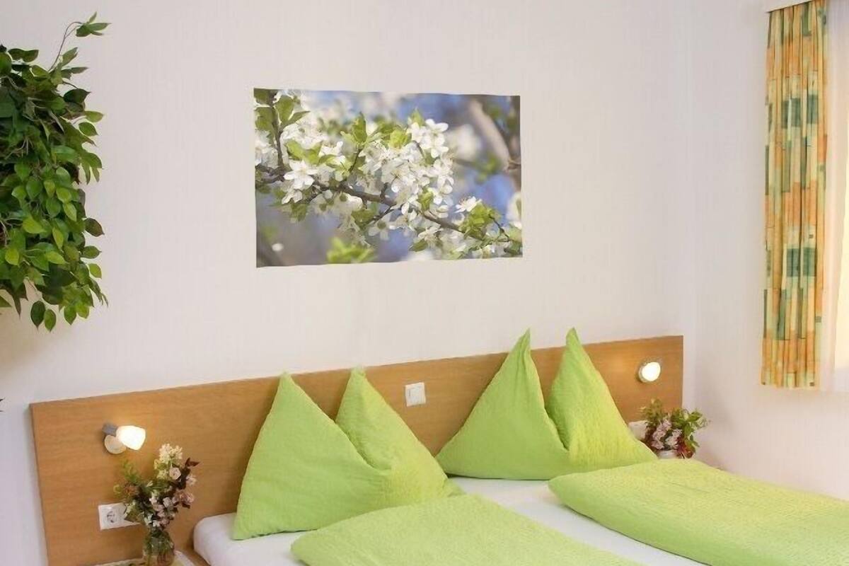 A bedroom with a wooden bed, green pillows, a nightstand with a lamp, and a wall-mounted picture.
