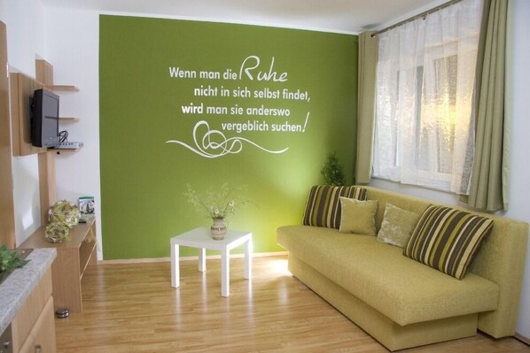 A living room with a green accent wall, a sofa, a small white table, and a television on a wooden stand. - Gasthof Spengerwirt (Hirschegg)