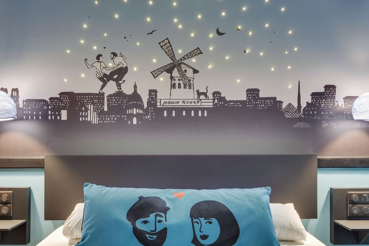 Ein Hotelzimmer mit Stadtbild-Wandgemälde, einem Bett mit Kopfkissen mit zwei Gesichtern und einem Nachttisch mit Lampe.