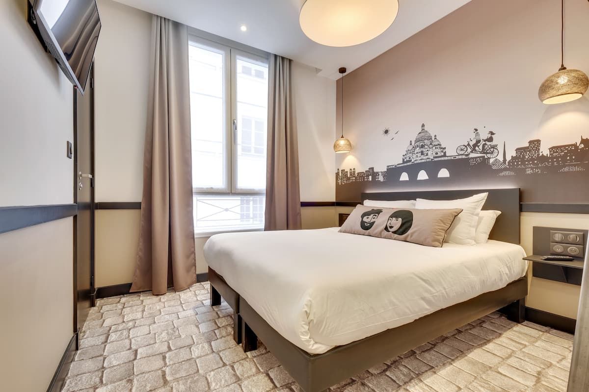 Ein Hotelzimmer mit einem großen Bett, einer Stadtkulissen-Mural an der Wand, einem Fenster mit Vorhängen und einem kleinen Tisch mit einer Lampe.