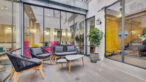 A modern outdoor seating area with wicker chairs and a small table, surrounded by glass walls and potted plants. - Hotel Lucien & Marinette (Paris)