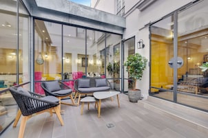A modern outdoor seating area with wicker chairs and a small table, surrounded by glass walls and potted plants.