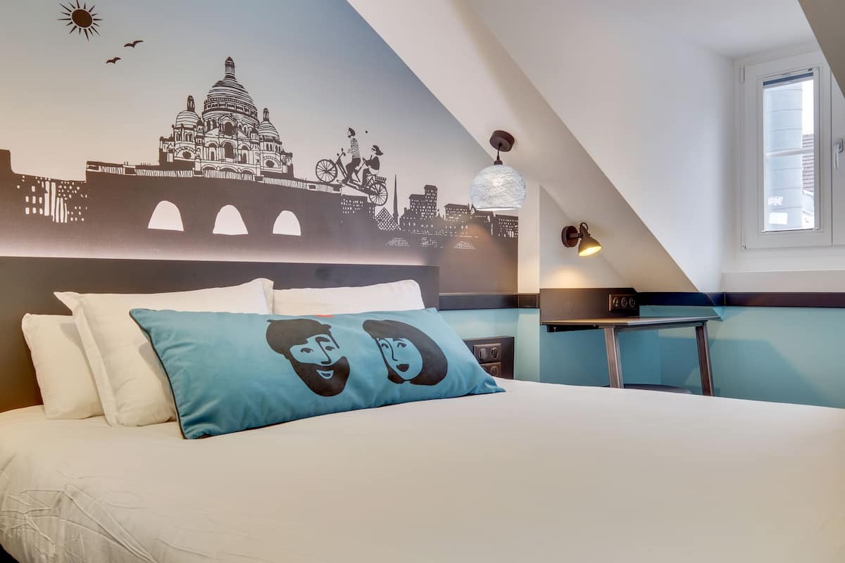 Ein Hotelzimmer mit einem Bett, einer Stadtansicht-Mural, einem Tisch mit Lampe und einem Fenster.