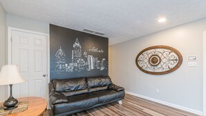 A room with a black leather sofa, a round wooden table with a lamp, a cityscape wall art, and a decorative metal piece on the wall. - Central Atlanta 8BR and 3600 sqft (Atlanta)