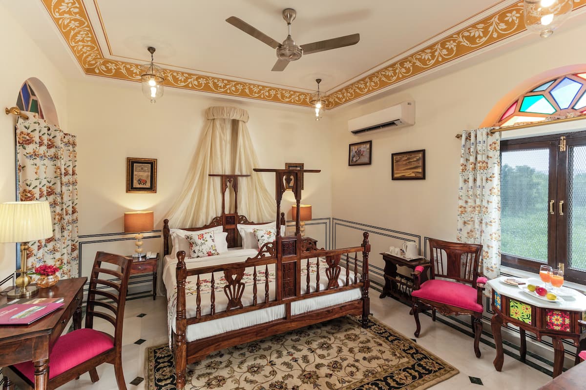 Un dormitorio con cama con dosel, ventilador de techo, candelabro, ventana con cortinas y una mesita con fruta y bebidas.