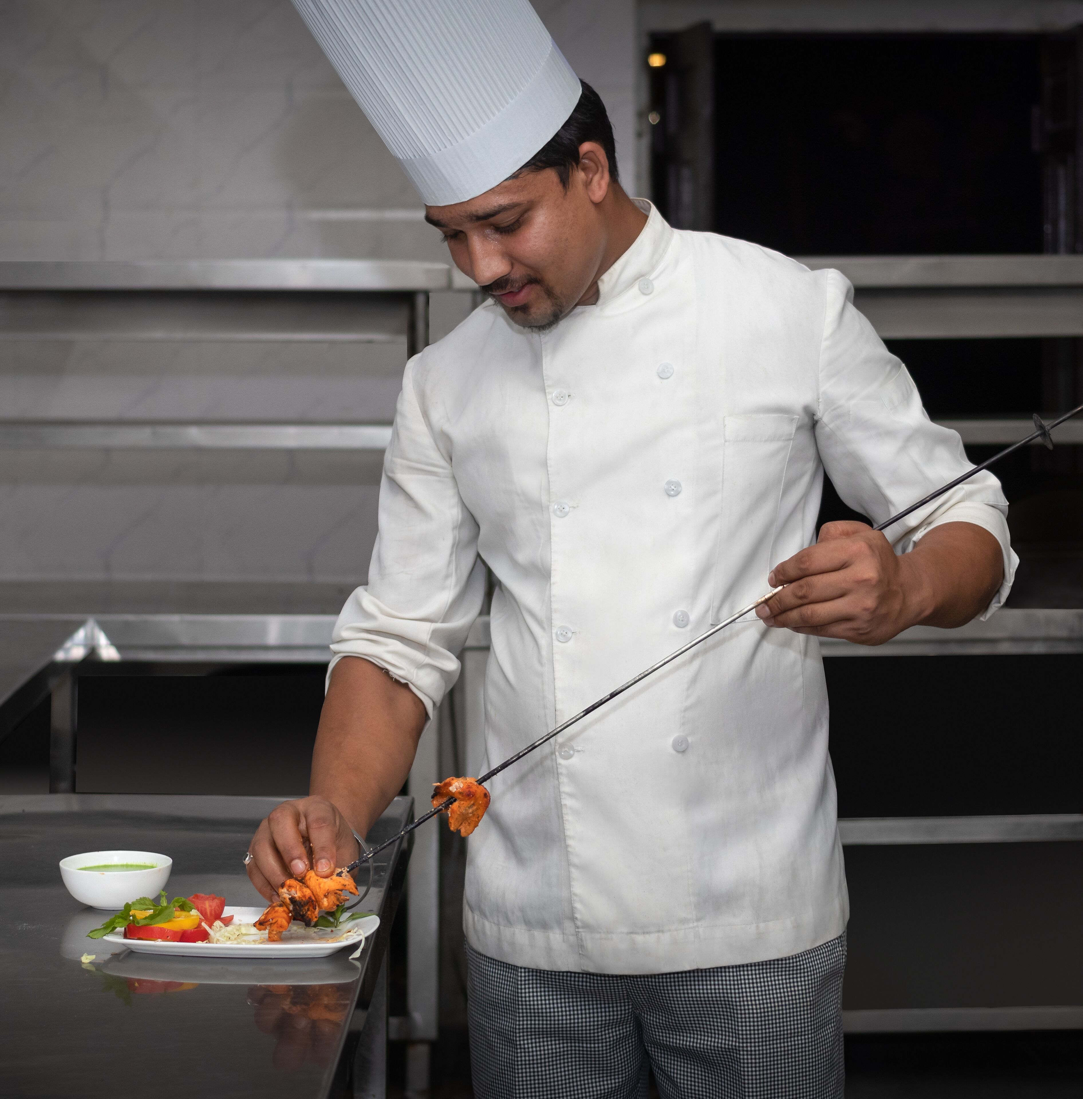 Un chef, vêtu d’une veste et d’un chapeau blancs, tient une pince au-dessus d’une assiette de nourriture.