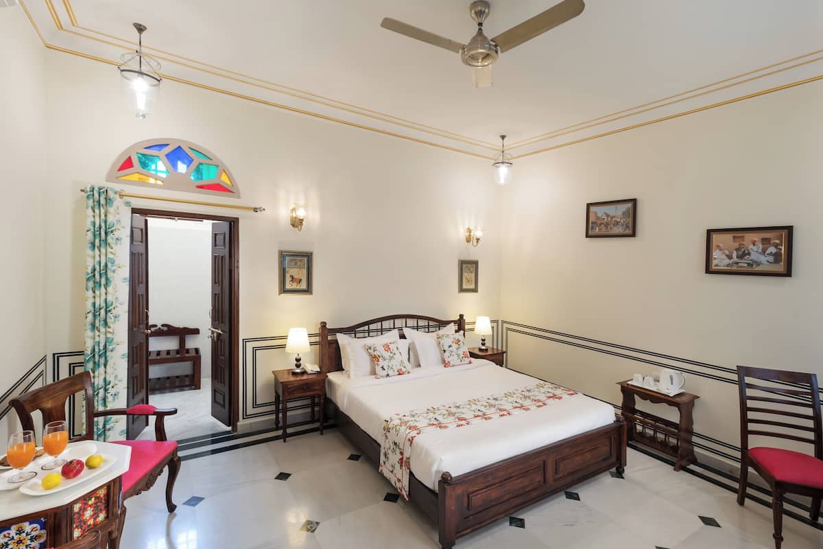 Habitación de hotel con cama, mesitas de noche, una silla, una mesa de comedor con fruta y bebidas, y un ventilador de techo.