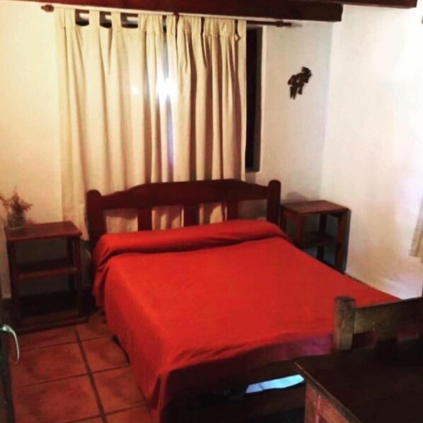 A bedroom with a bed, a nightstand, a small table, and a window with curtains. - Cabañas Altos de Tafi (El Mollar)