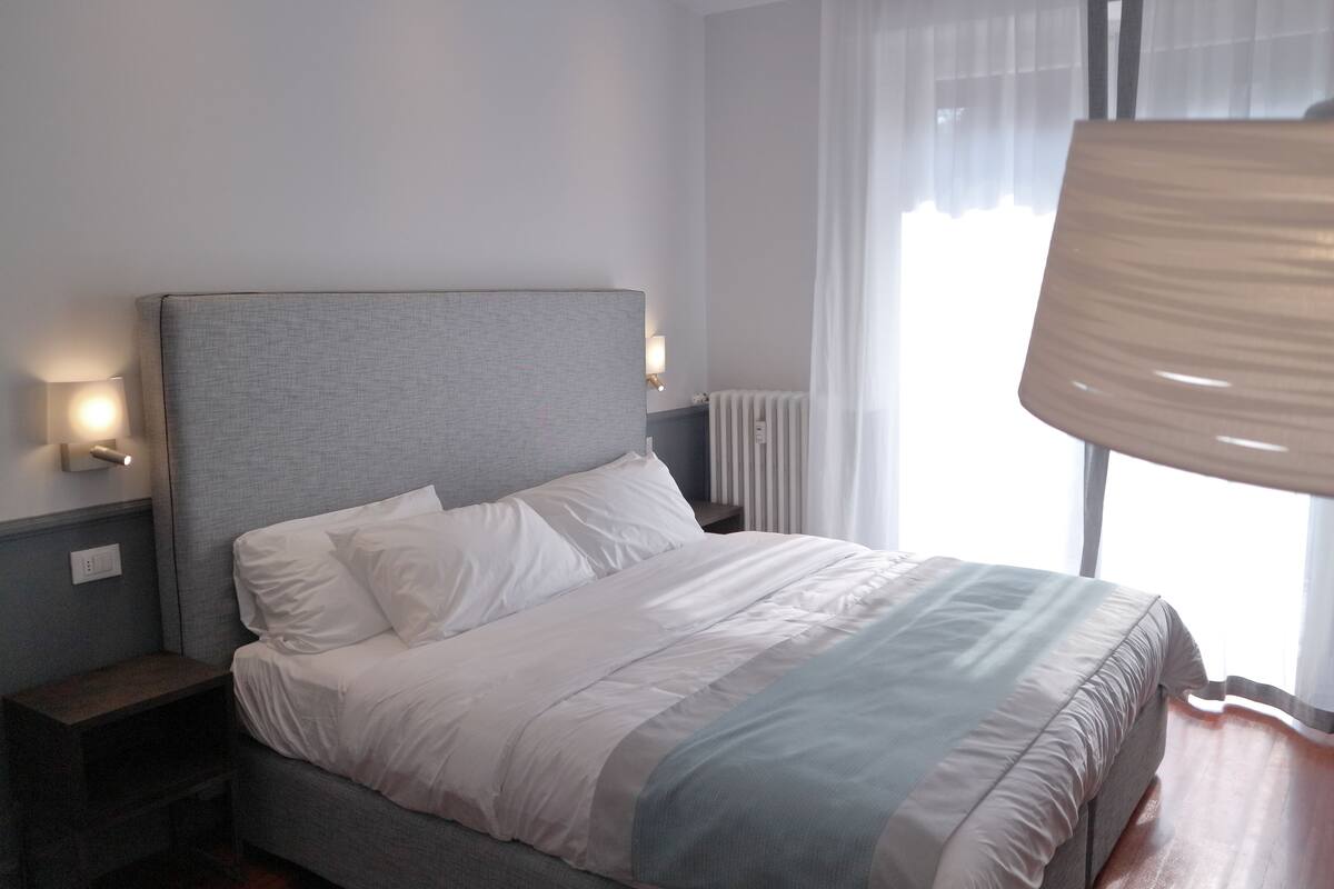 A neatly made bed with white linens and a grey headboard, a bedside table with a lamp, and a window with sheer curtains.