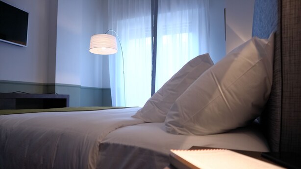 A neatly made bed with white linens, a bedside table with a lamp, and a window with sheer curtains. - Dimora dell'Arte B&B (Sesto San Giovanni)