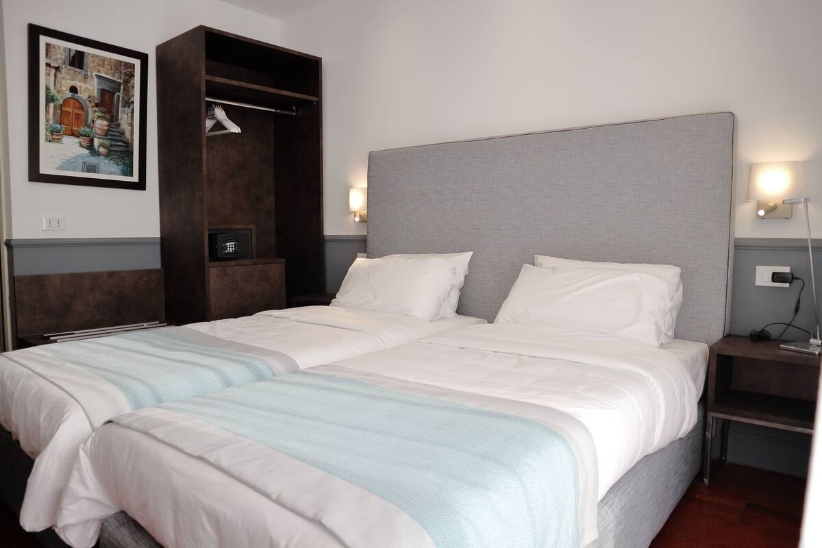 A double bed with a grey headboard, white bedding, and a light blue throw blanket. There is a wooden wardrobe with a mirror and a framed picture on the wall.