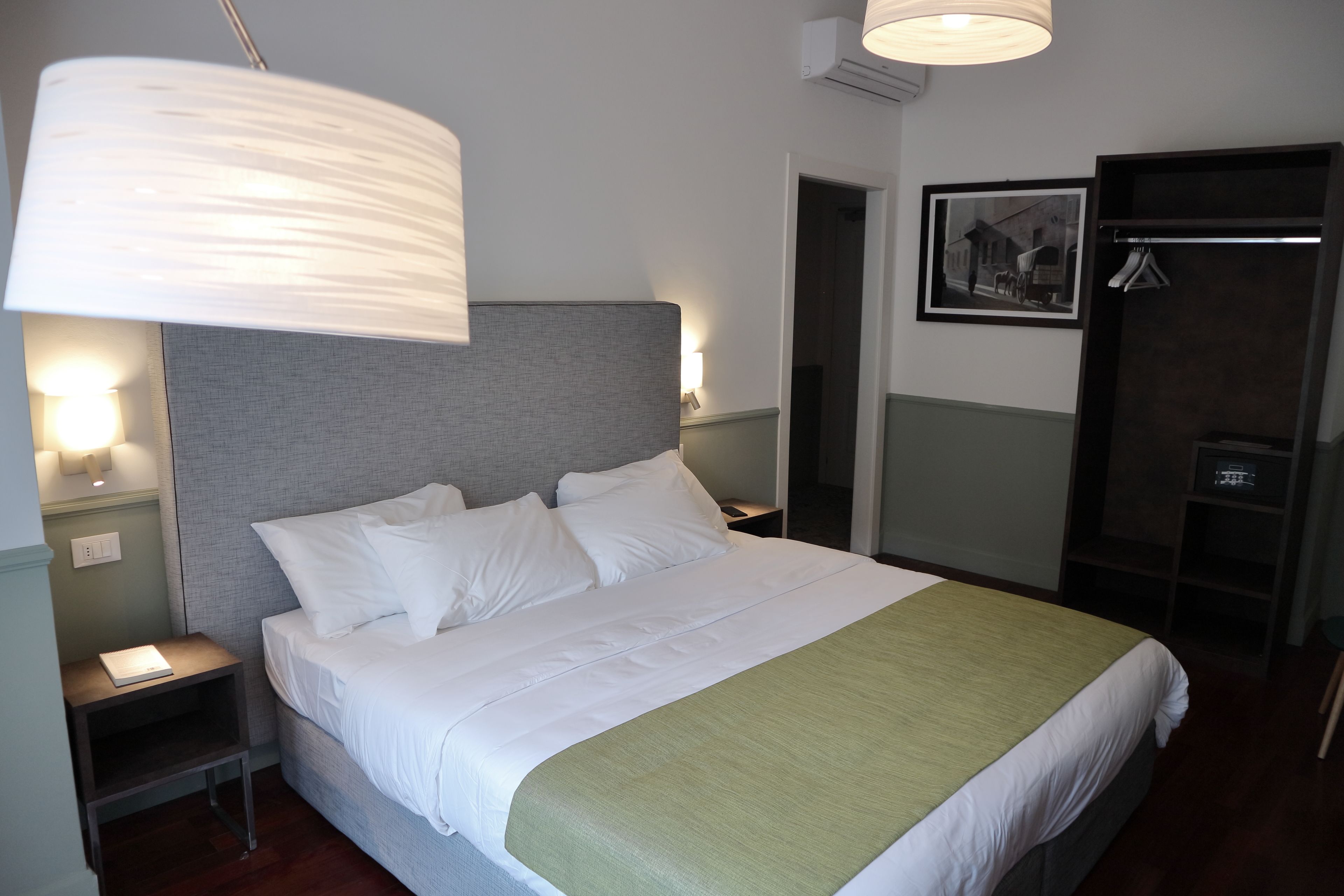 Uma cama bem arrumada com lençóis brancos e um cobertor verde, um abajur e um quadro na parede.