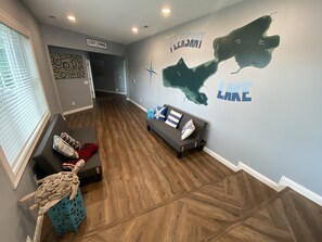 A spacious living area with a wooden floor, a gray sofa with decorative pillows, and a wall mural depicting a map and the words "Come be PLEASANT LAKE."