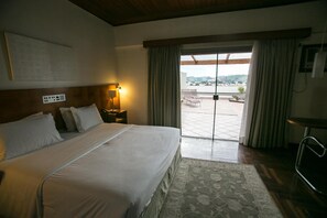 A hotel room with a large bed, a bedside table with a lamp, a view of the outdoors through a glass door, and a small table with chairs.