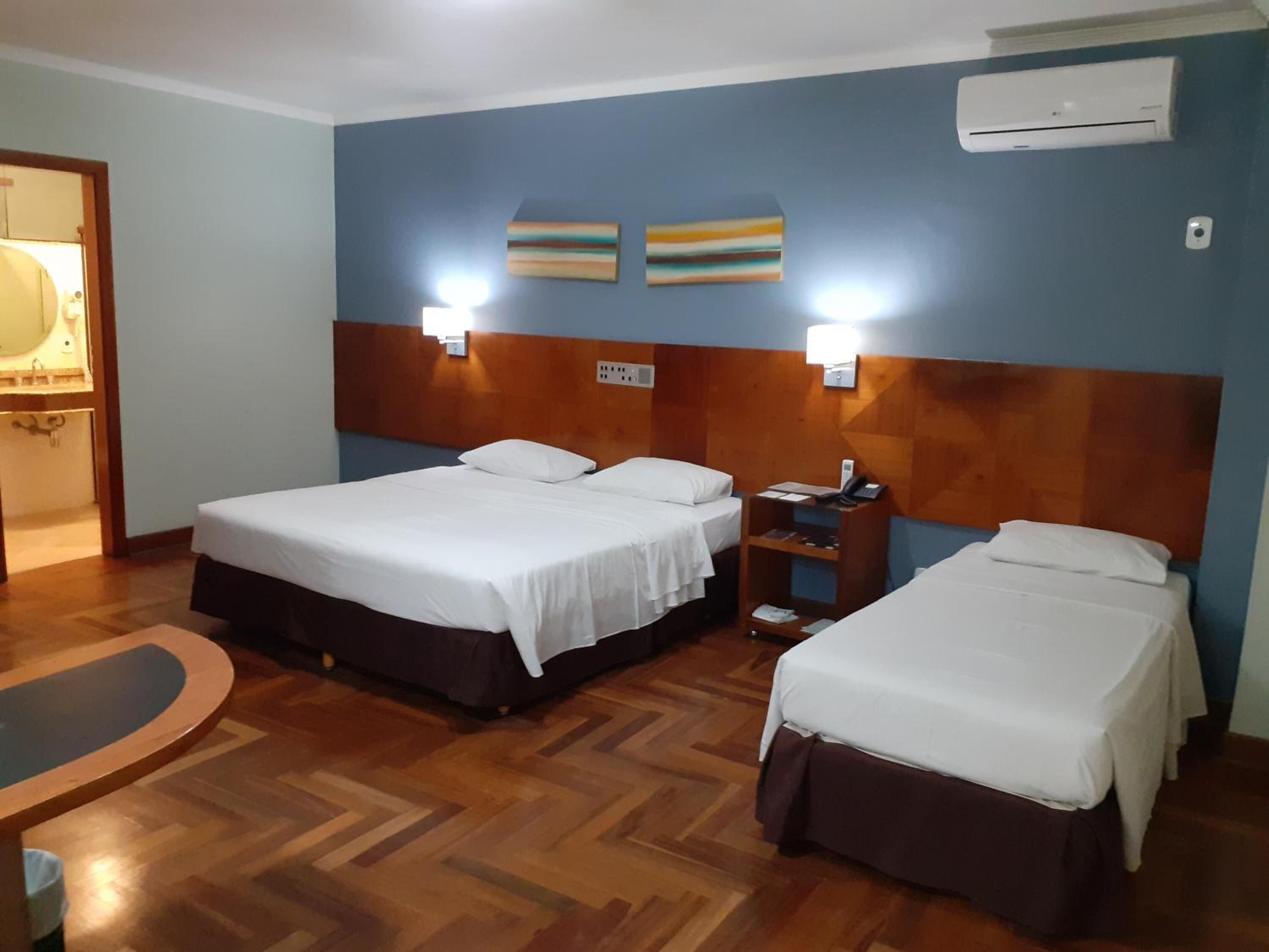 A hotel room with two beds, a wooden floor, a desk with a chair, and an air conditioning unit.