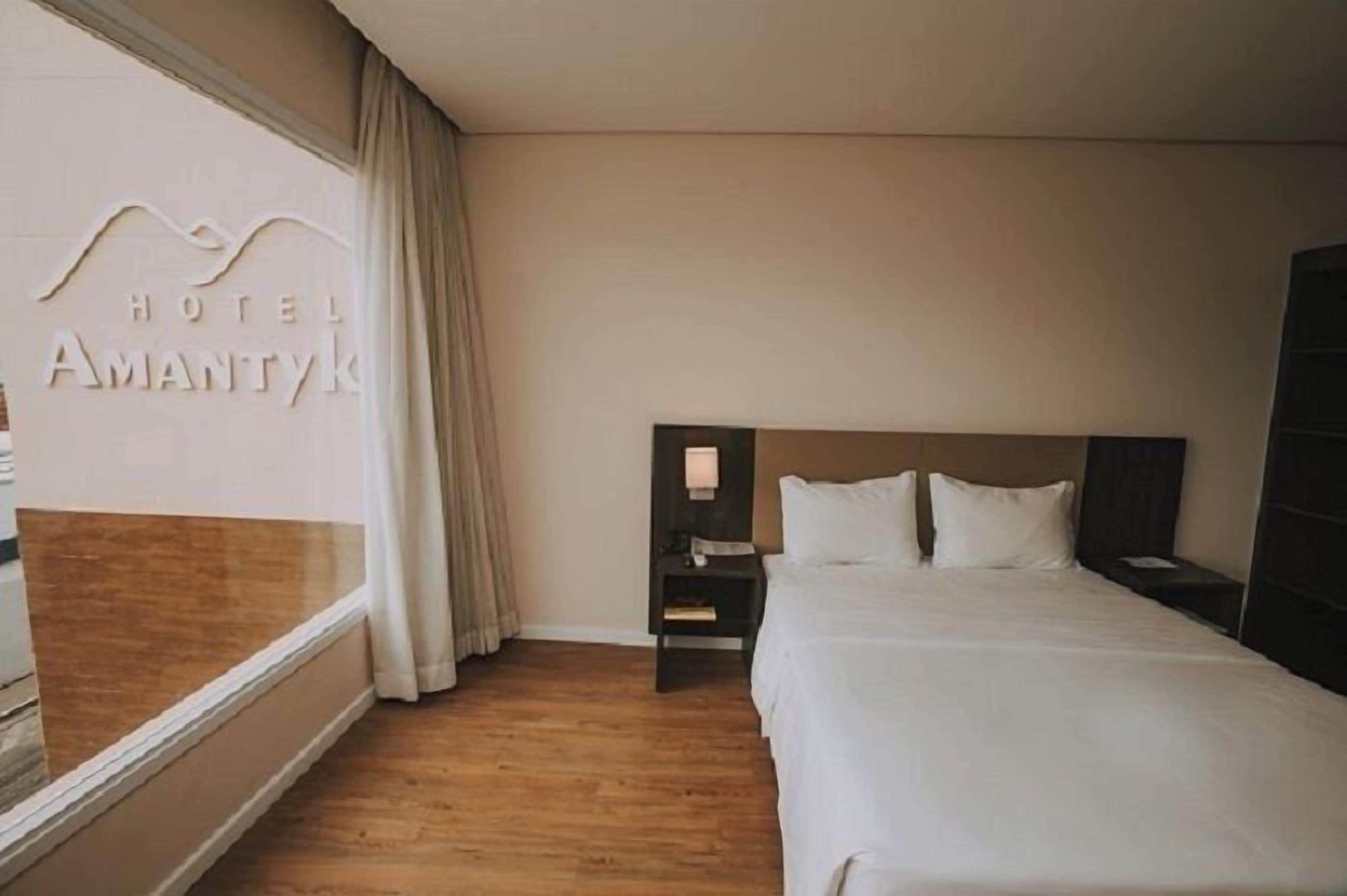 Habitación de hotel con un ventanal grande, una cama con ropa blanca, una mesita de noche con lámpara y suelo de madera.