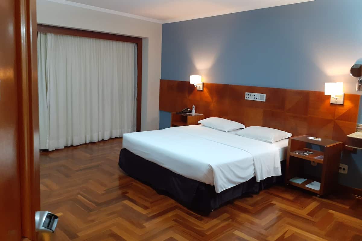 Ein Hotelzimmer mit Bett, Holzboden, Vorhängen, einem Nachttisch und einer Wandlampe.