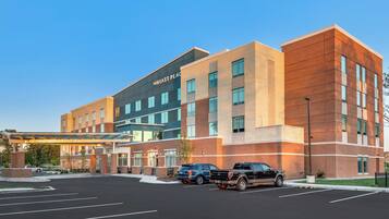 A modern hotel with a glass entrance, a covered walkway, and a parking lot with several vehicles.