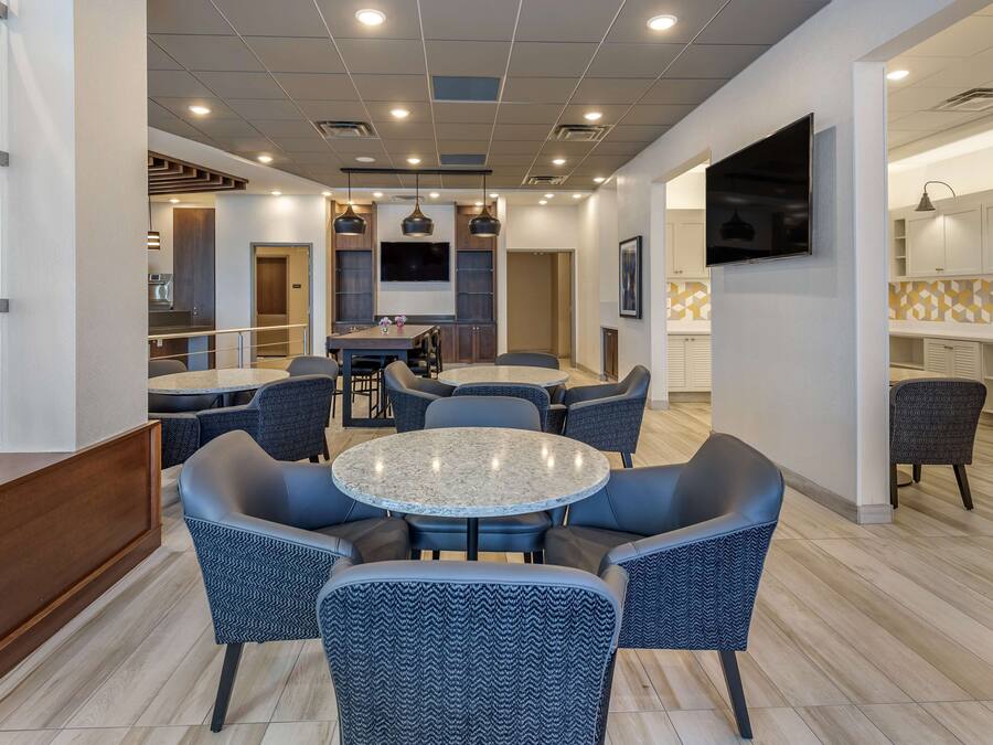 A modern hotel lobby with a round dining table surrounded by chairs, a kitchen area in the background, and a flat-screen TV mounted on the wall.