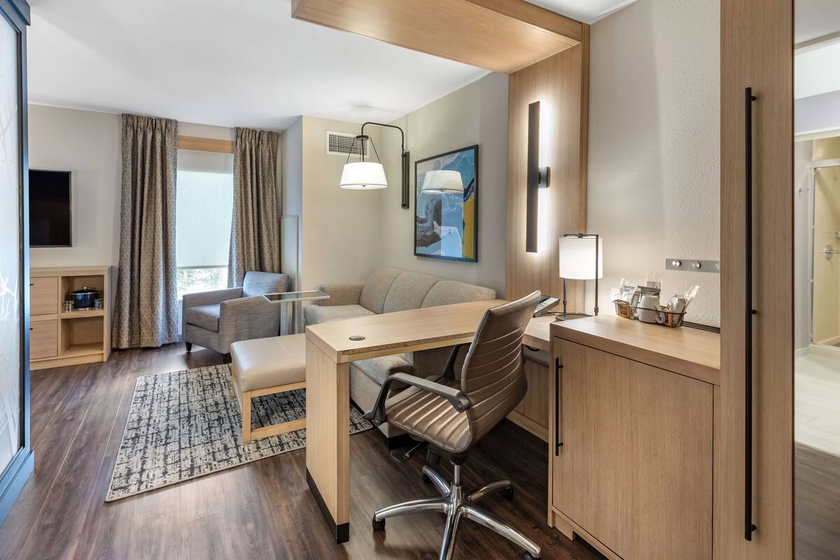 A modern hotel room with a wooden desk, a cushioned chair, a sofa, and a flat-screen TV.