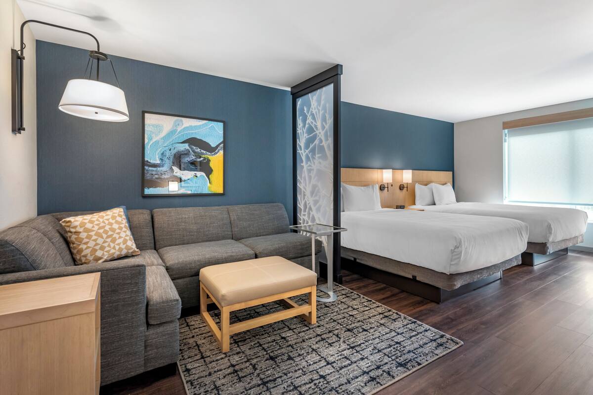 A modern hotel room with a grey sofa, a wooden bench, a bed with two white bedsheets, and a large window.