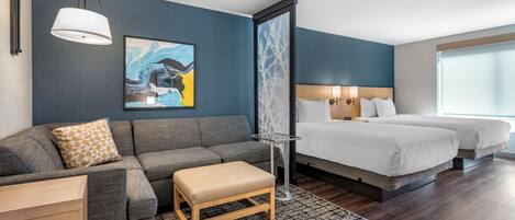 Una habitación de hotel moderna con un sofá gris, un banco de madera, una cama con dos sábanas blancas y un ventanal grande.