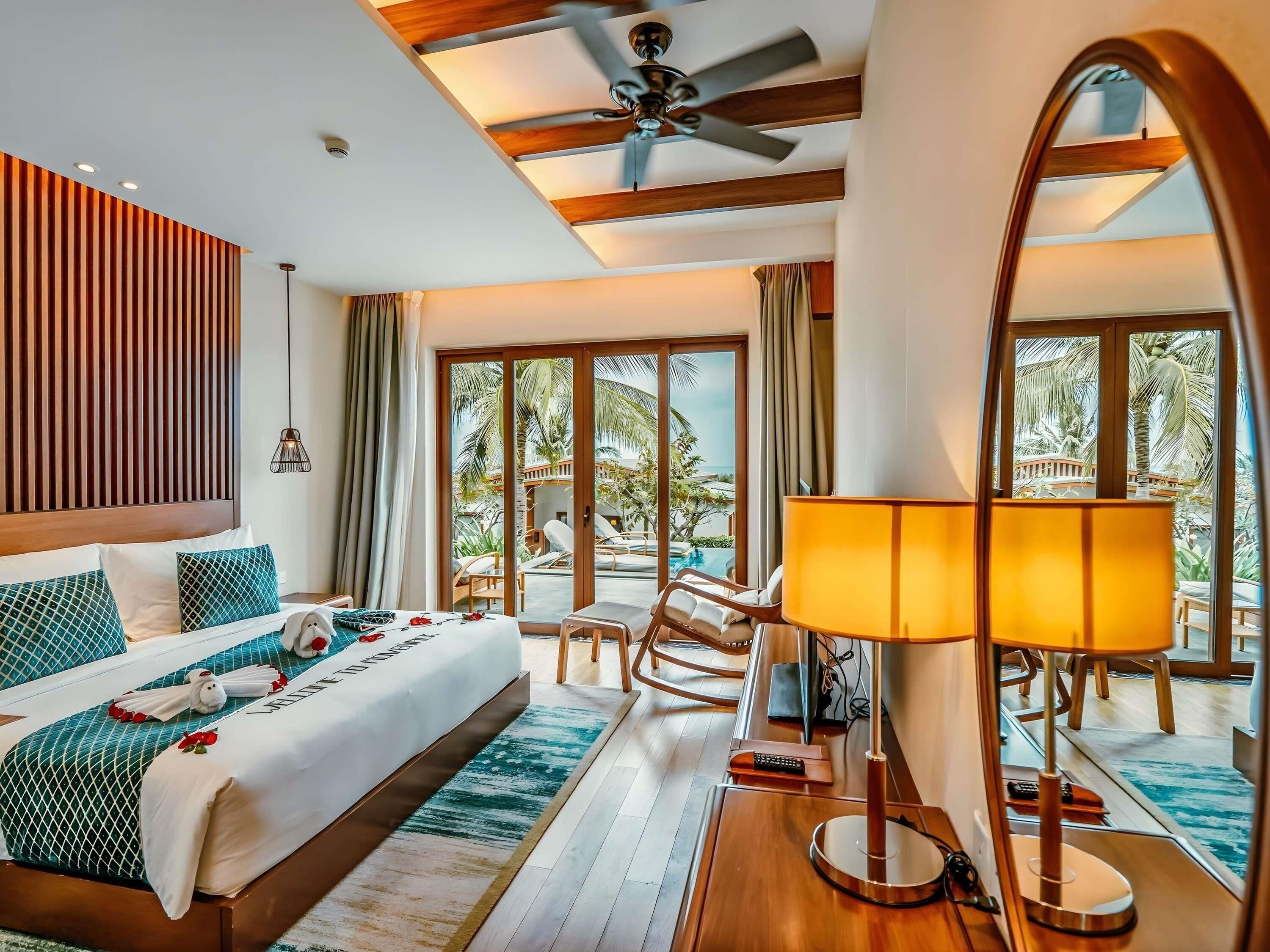 A hotel room with a bed, a desk, a chair, a lamp, and a view of a pool and palm trees.