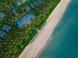 An aerial view of a coastal resort with multiple villas, a sandy beach, and a clear blue ocean.