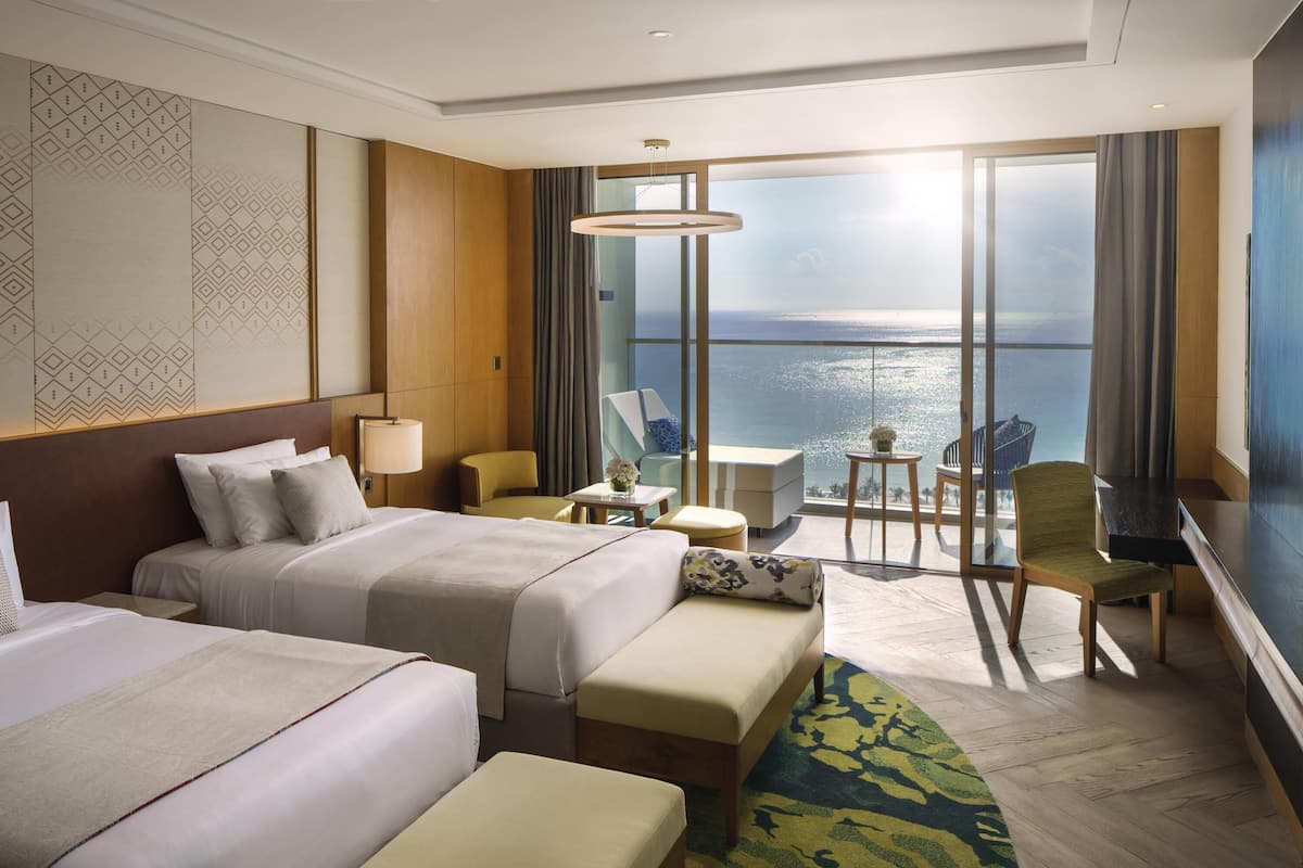 Habitación de hotel con dos camas, un escritorio, una silla, una mesita y vistas al mar.